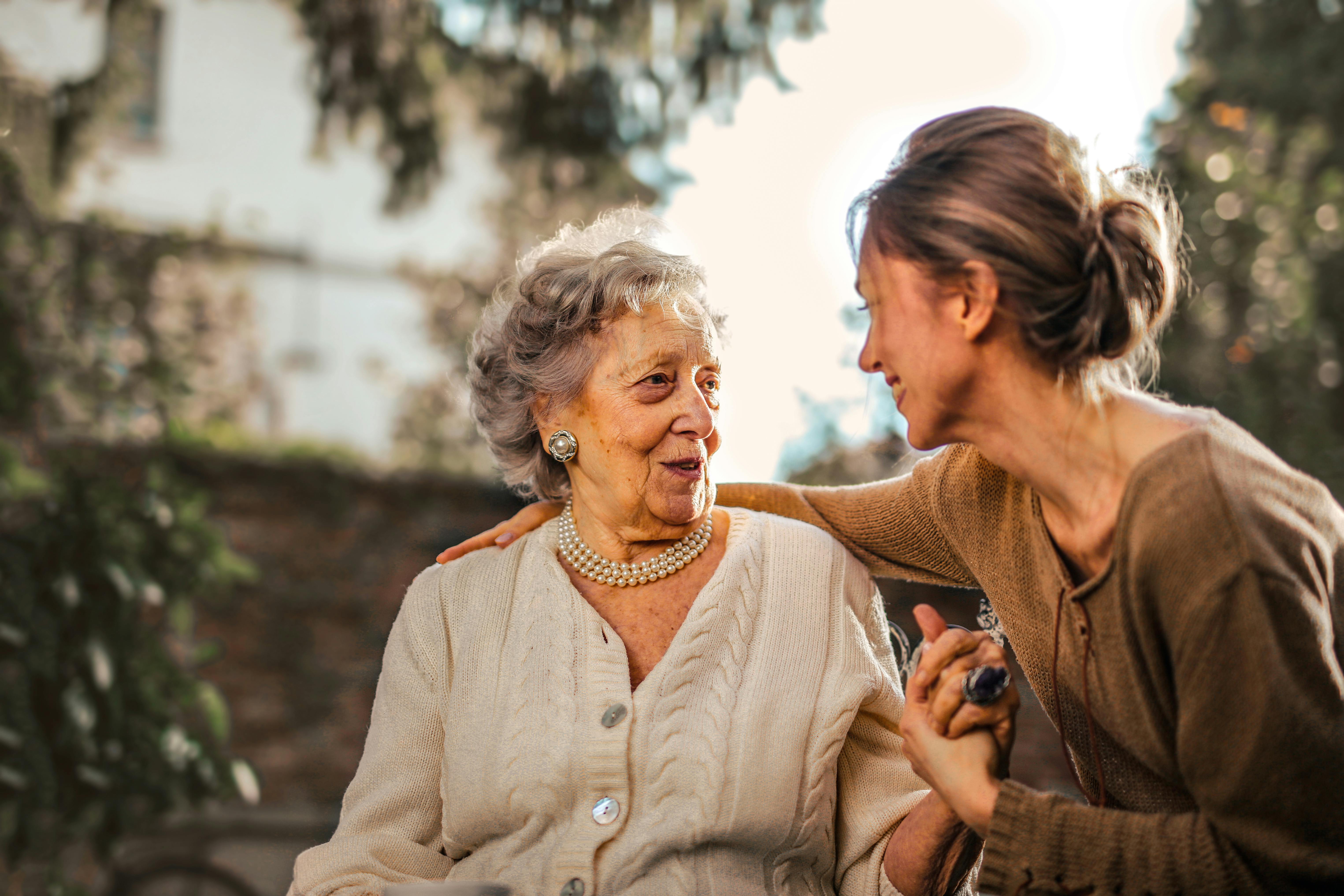 An older daughter holds her elderly mothers' hand as they chat on a bench. 