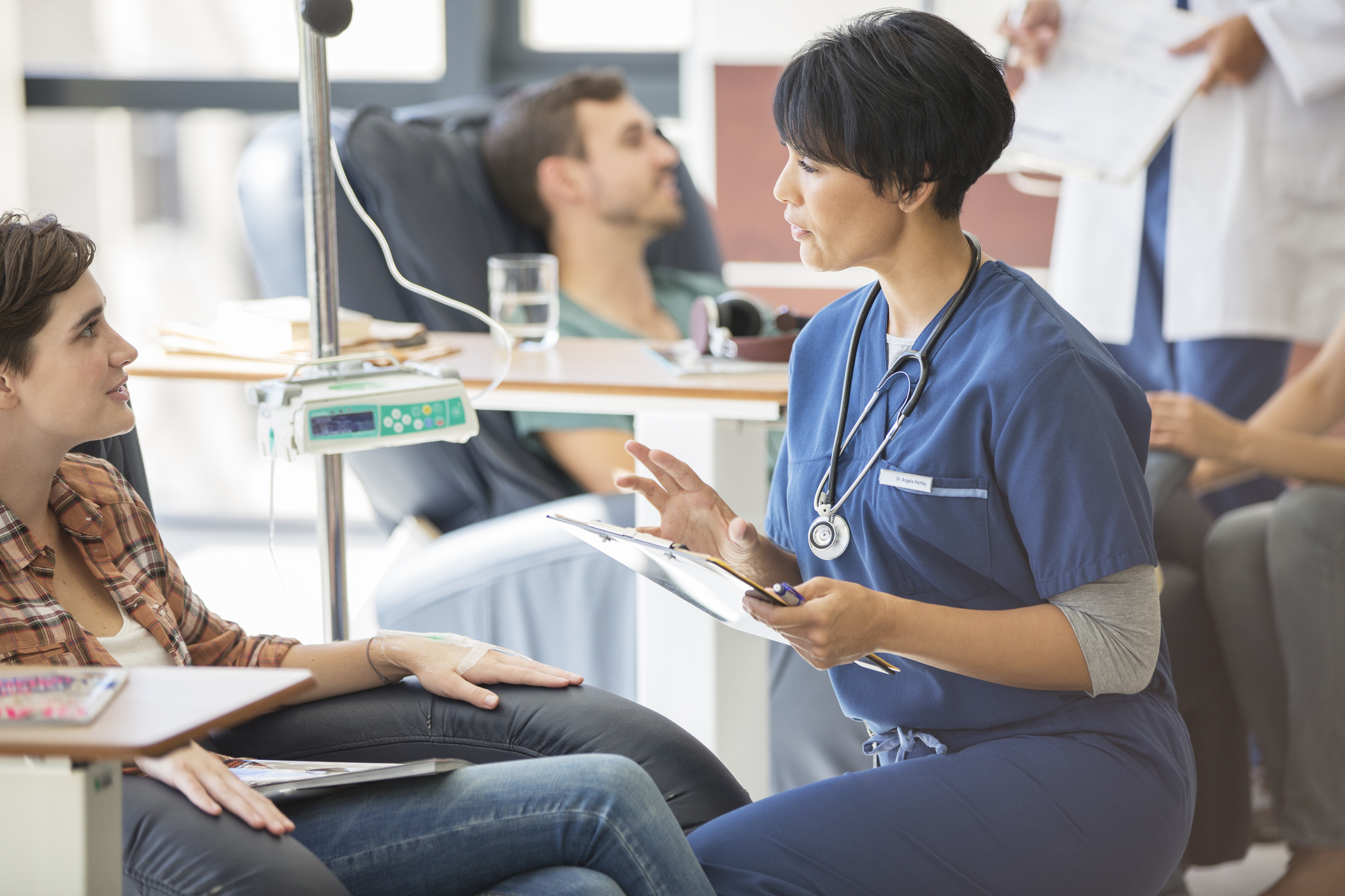 Depiction of a nurse treating a patient in a clinical trial. Patient is sitting facing the nurse with an IV bag next to them.
