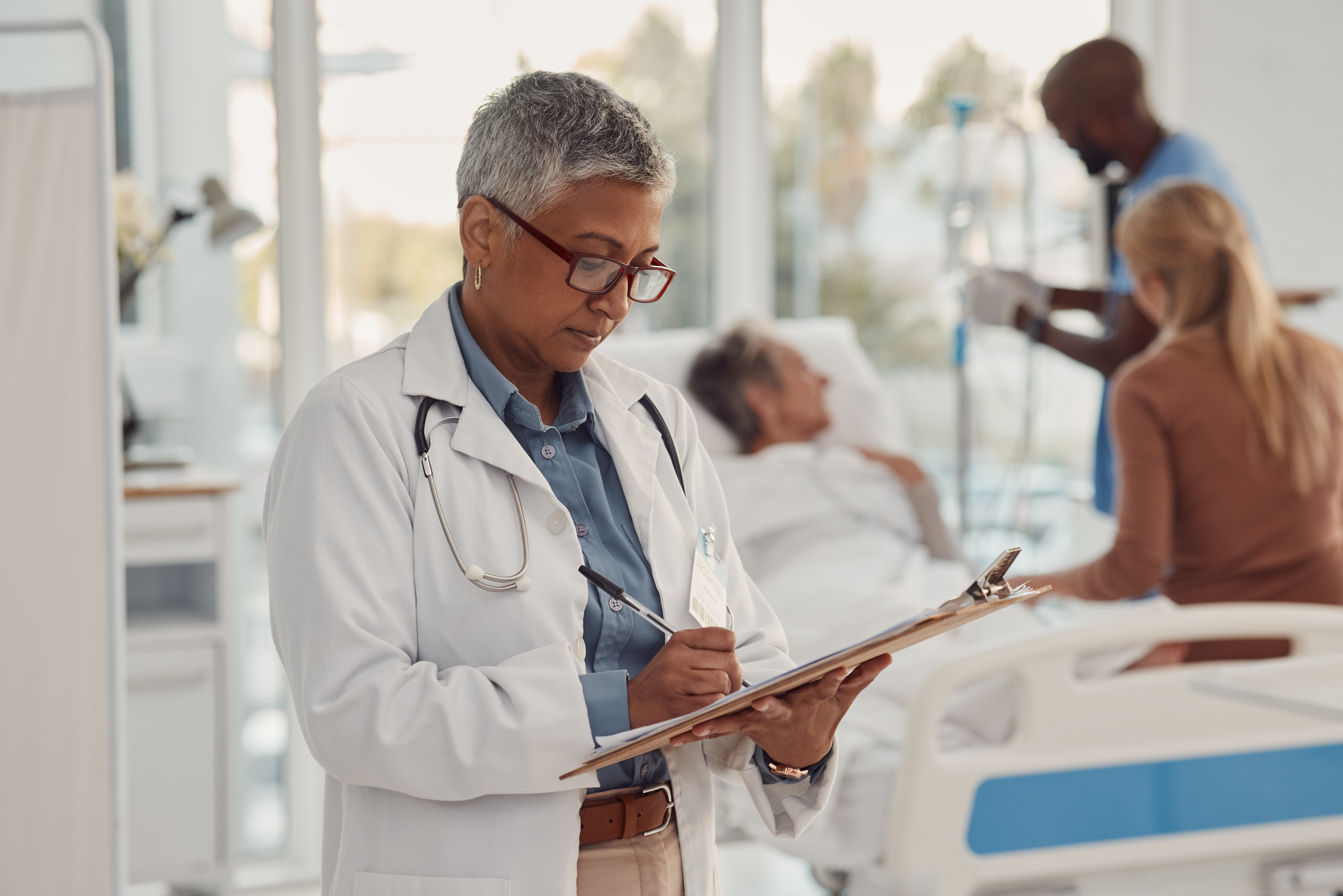 A healthcare worker refers to a chart while another attends to a patient in the background.