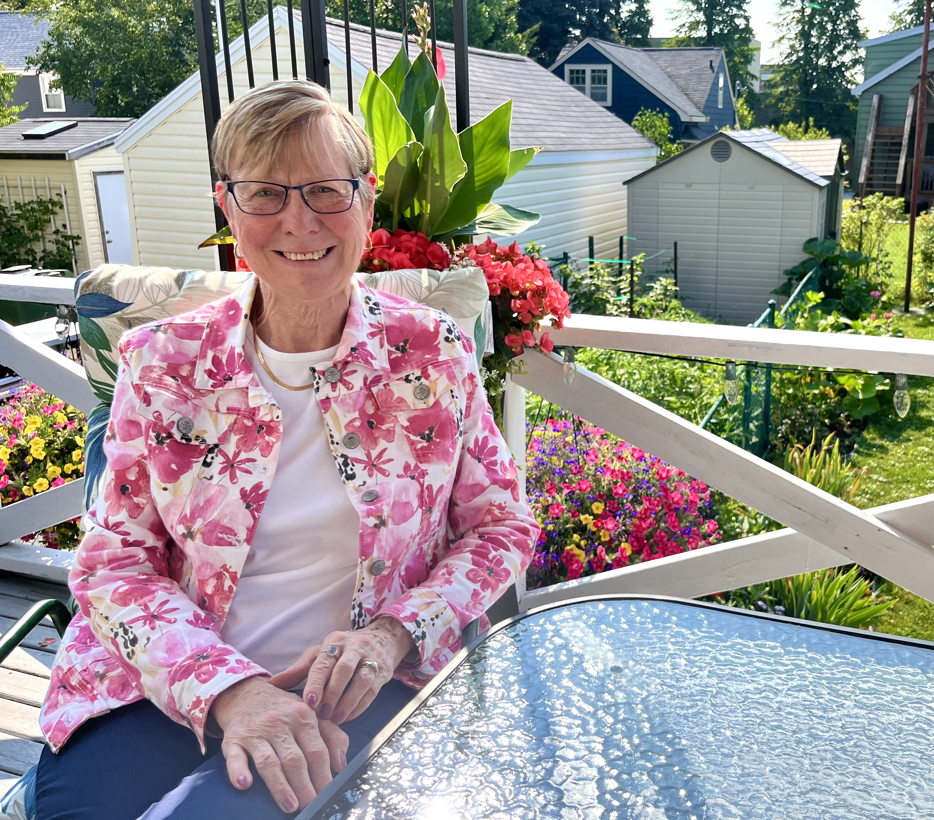 Debbie Purchase smiles at the camera wearing a pink and white floral jacket in her sunny backyard. 
