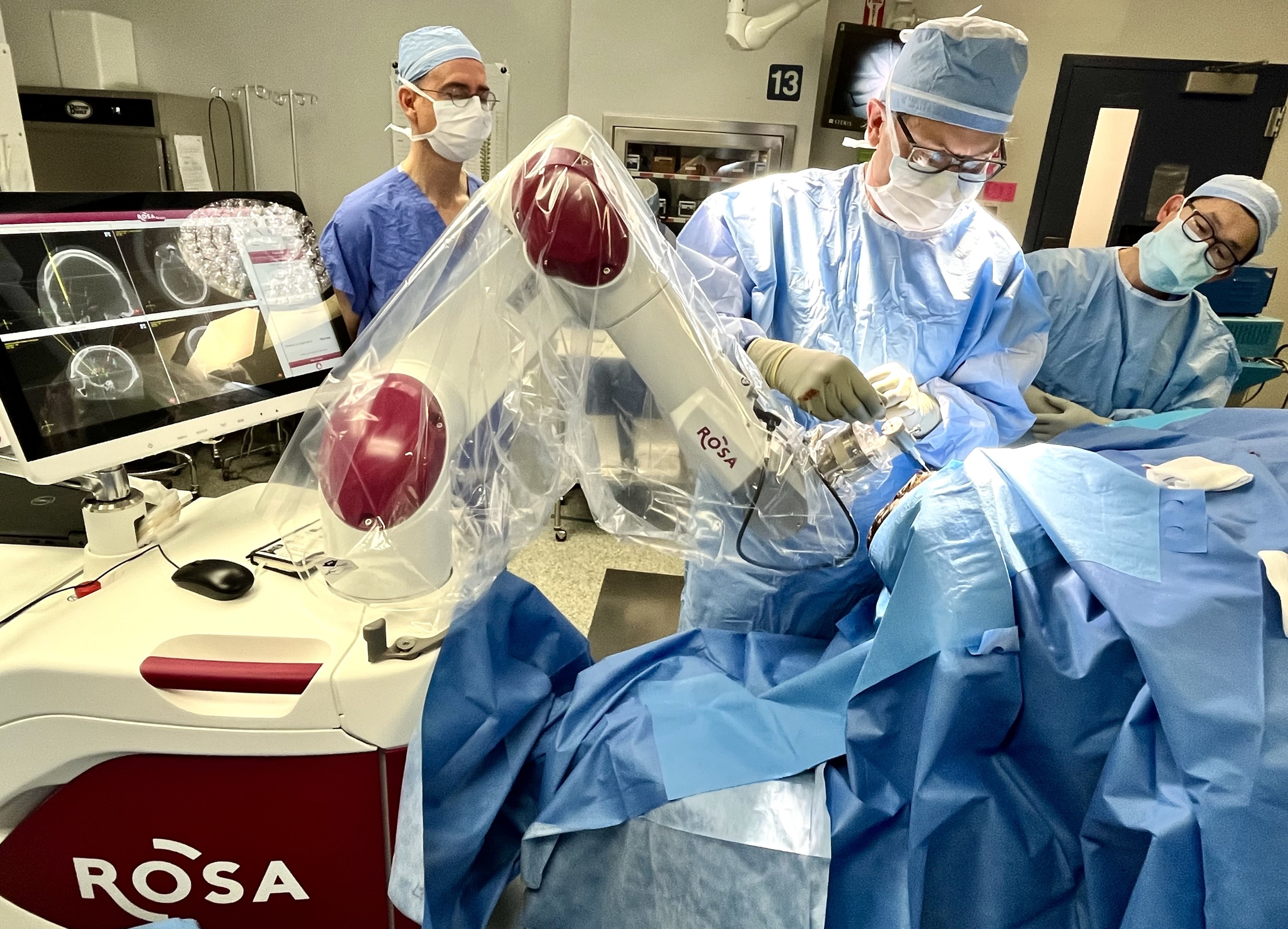 A surgeon wearing blue scrubs and a blue face mask operates on a patient's brain using a surgical robot. Two other surgeons dressed in blue scrubs with blue masks look on from behind him.
