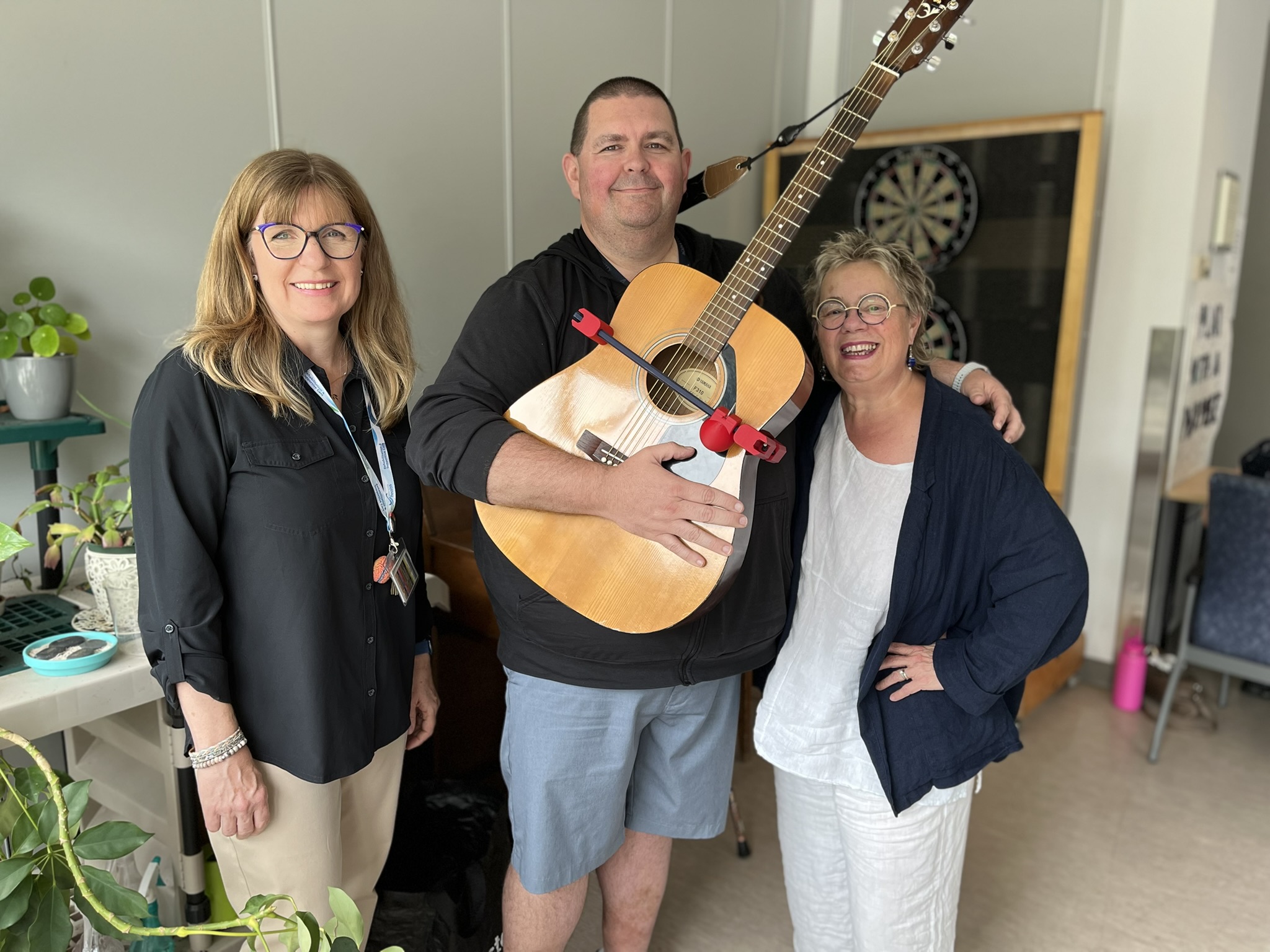 Picture of three Nova Scotia Health employees, two female and one male, with the male holding a guitar fitted with an assistive 3D-printed device called StrumAssist.