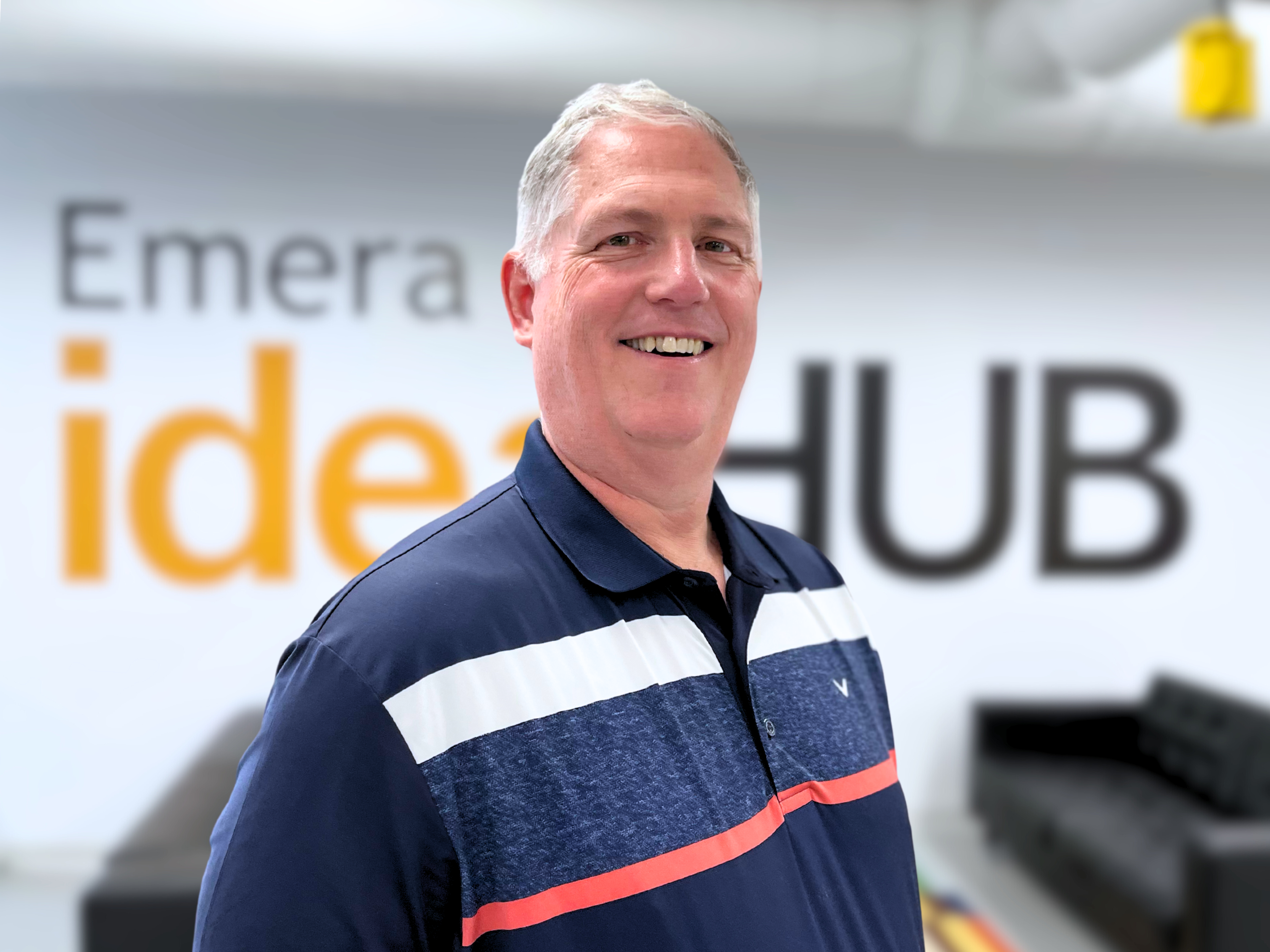 A photo of Dr. Clif Johnston, a man with grey hair wearing a blue, white and red polo shirt, smiling at the camera in front of a wall that says "Emera IdeaHub"
