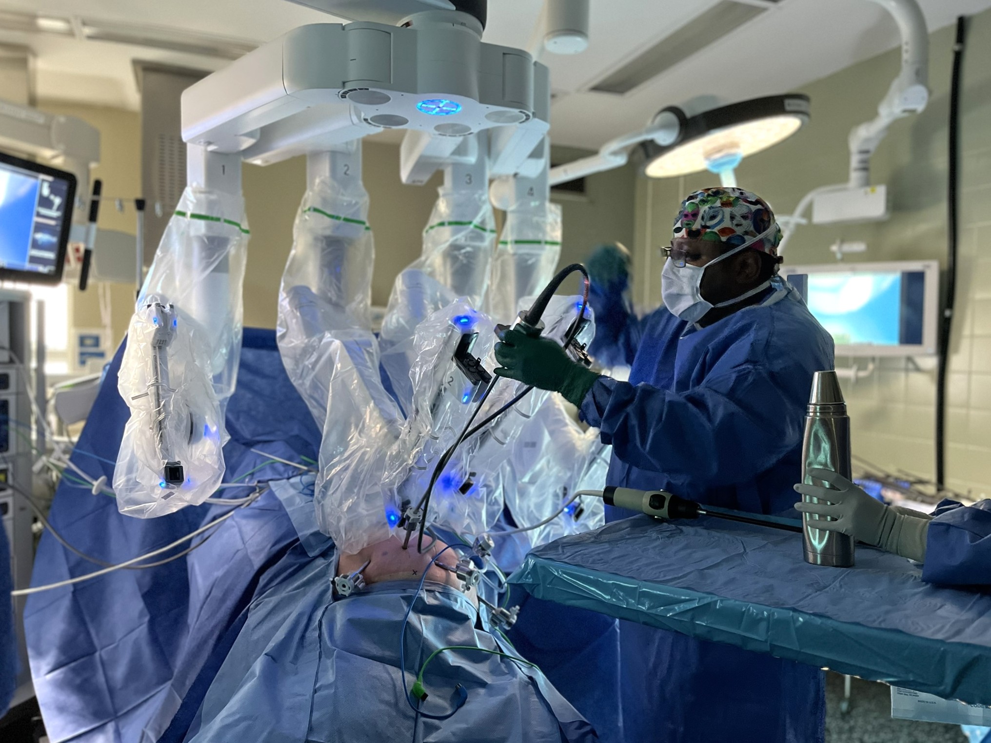 A large surgical robot with 4 long arms placed over a patient on the operating table in an OR, with a surgeon attaching instruments to it. 