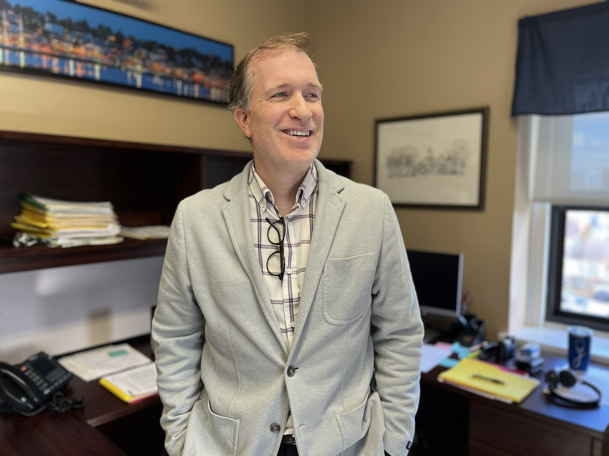 A smiling man standing in an office with hands in pockets wearing a grey blazer with glasses tucked into his collar. The man pictured is named Dr. Ashley Davidson.