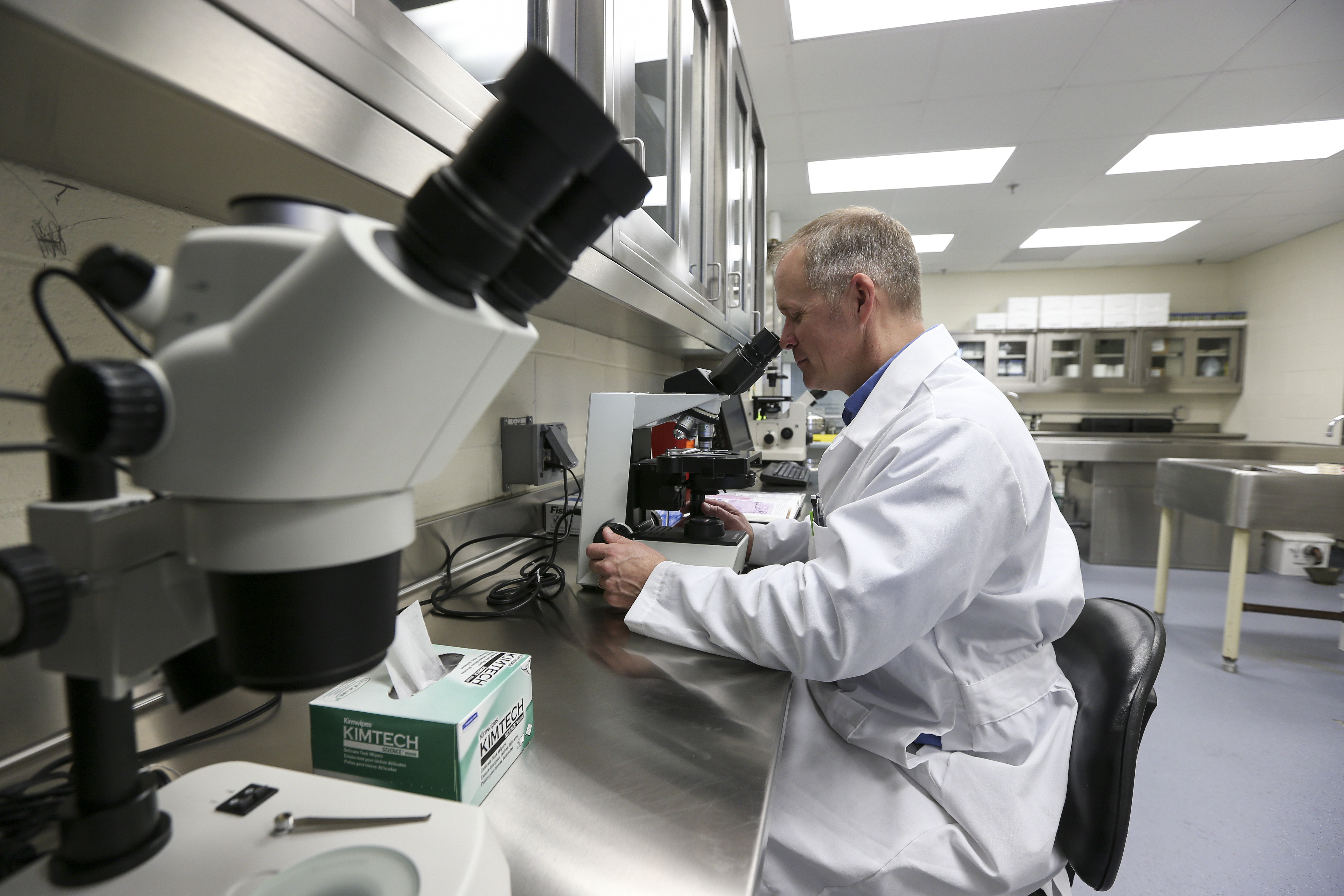 A man wearing a white lab coat in a lab looking into a microscope. 