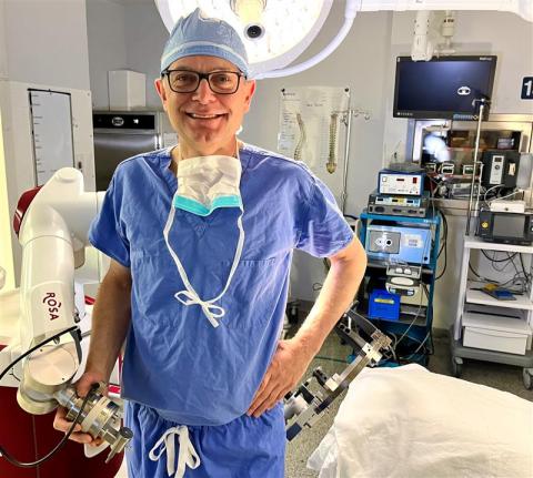 Dr. David Clarke poses with the ROSA ONE surgical robot in an operating room. He is wearing blue scrubs, glasses and a mask is hanging around his neck.