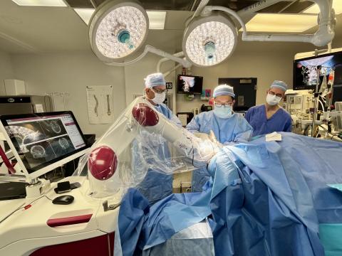 Large, circular lights are seen behind three surgeons wearing blue scrubs operate on a patient's brain with the help of a surgical robot. 