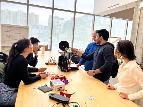 Four students and their instructor sit at a large workbench working with a 3D printer in a brightly-lit room with massive windows behind them. 