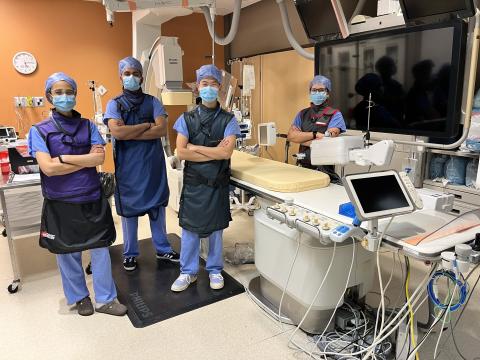 Four students wearing scrub, masks and lead vests stand with their arms crossed across their chests in a cardiac catheterization lab. A hospital bed, equipment and wires are seen all around them. 