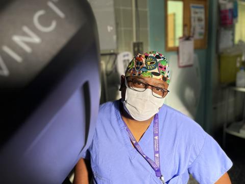 A male surgeon wearing a scrub, a mask, and a colorful cap is sitting at the console of a surgical robot, which can be partially seen on the left side of the image. 