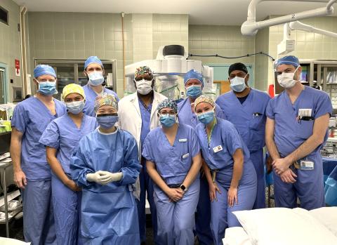 A group of 10 clinicians in an operating room wearing scrubs, mask, and hats. They are standing in front of a surgical robot, which can be partially seen behind and above them.
