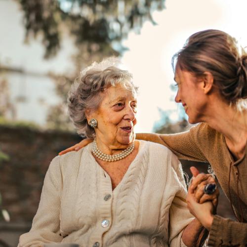 An older daughter holds her elderly mothers' hand as they chat on a bench. 