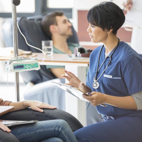 Depiction of a nurse treating a patient in a clinical trial. Patient is sitting facing the nurse with an IV bag next to them.