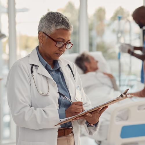 A healthcare worker refers to a chart while another attends to a patient in the background.