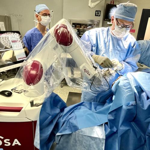 A surgeon wearing blue scrubs and a blue face mask operates on a patient's brain using a surgical robot. Two other surgeons dressed in blue scrubs with blue masks look on from behind him.