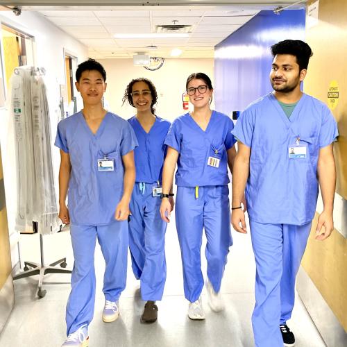 Four students wearing blue scrubs walk through double doors in a hospital hallway. 