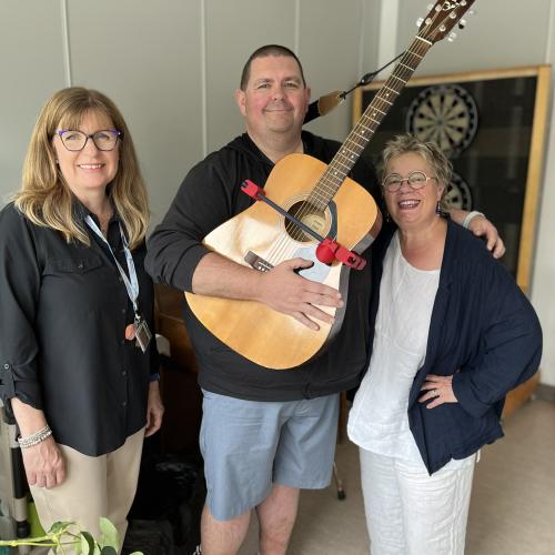 Picture of three Nova Scotia Health employees, two female and one male, with the male holding a guitar fitted with an assistive 3D-printed device called StrumAssist.