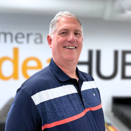 A photo of Dr. Clif Johnston, a man with grey hair wearing a blue, white and red polo shirt, smiling at the camera in front of a wall that says "Emera IdeaHub"