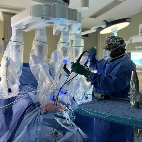A large surgical robot with 4 long arms placed over a patient on the operating table in an OR, with a surgeon attaching instruments to it. 