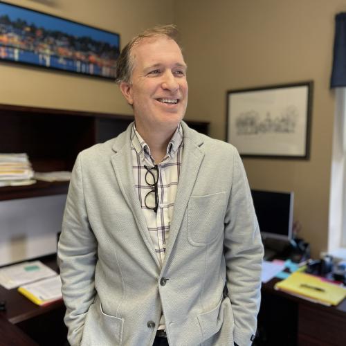 A smiling man standing in an office with hands in pockets wearing a grey blazer with glasses tucked into his collar. The man pictured is named Dr. Ashley Davidson.