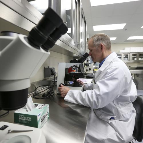 A man wearing a white lab coat in a lab looking into a microscope. 